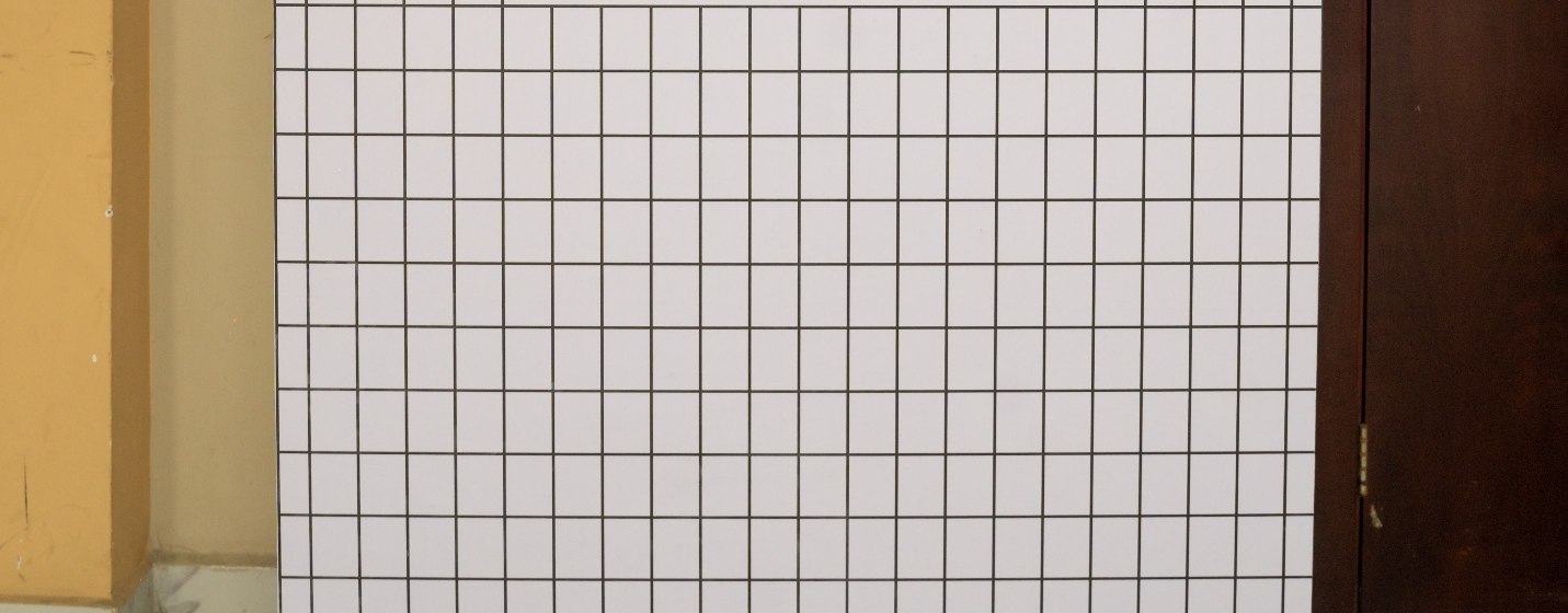 A detail of the gridded wall of the Losing Daylight exhibition at Glover Memorial Hall — white panels ruled into squares, between a painted pier and a dark timber door.