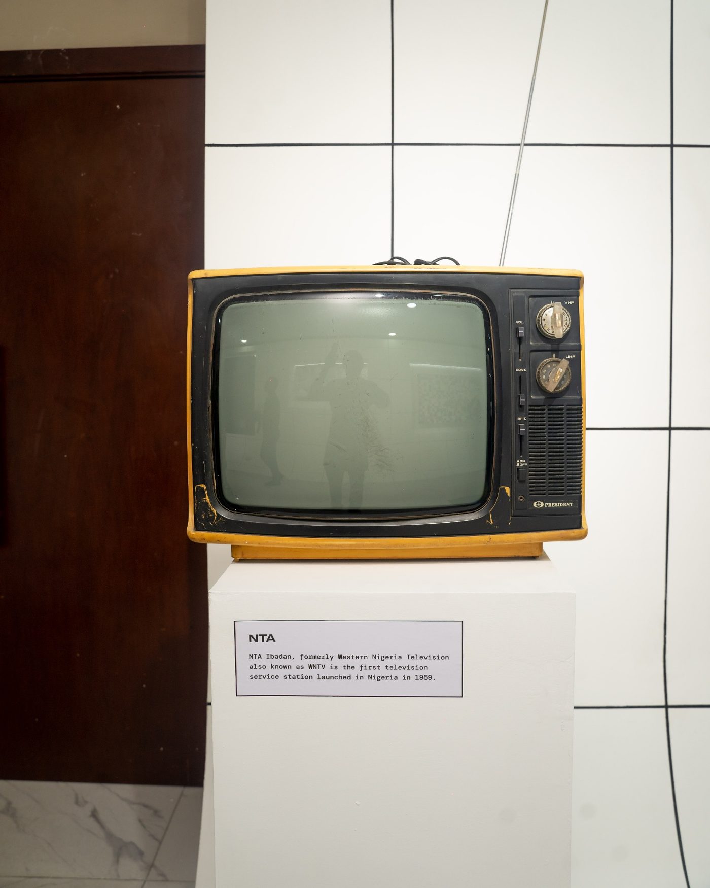 A vintage yellow-cased NTA television set on a white plinth, with its caption card below.