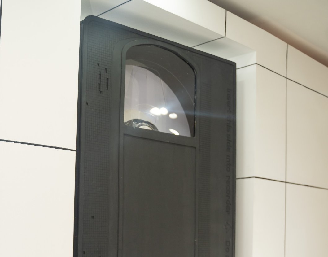 An oversized VHS cassette mounted in a recessed vitrine in the white tiled gallery wall, its spool window catching the ceiling lights.